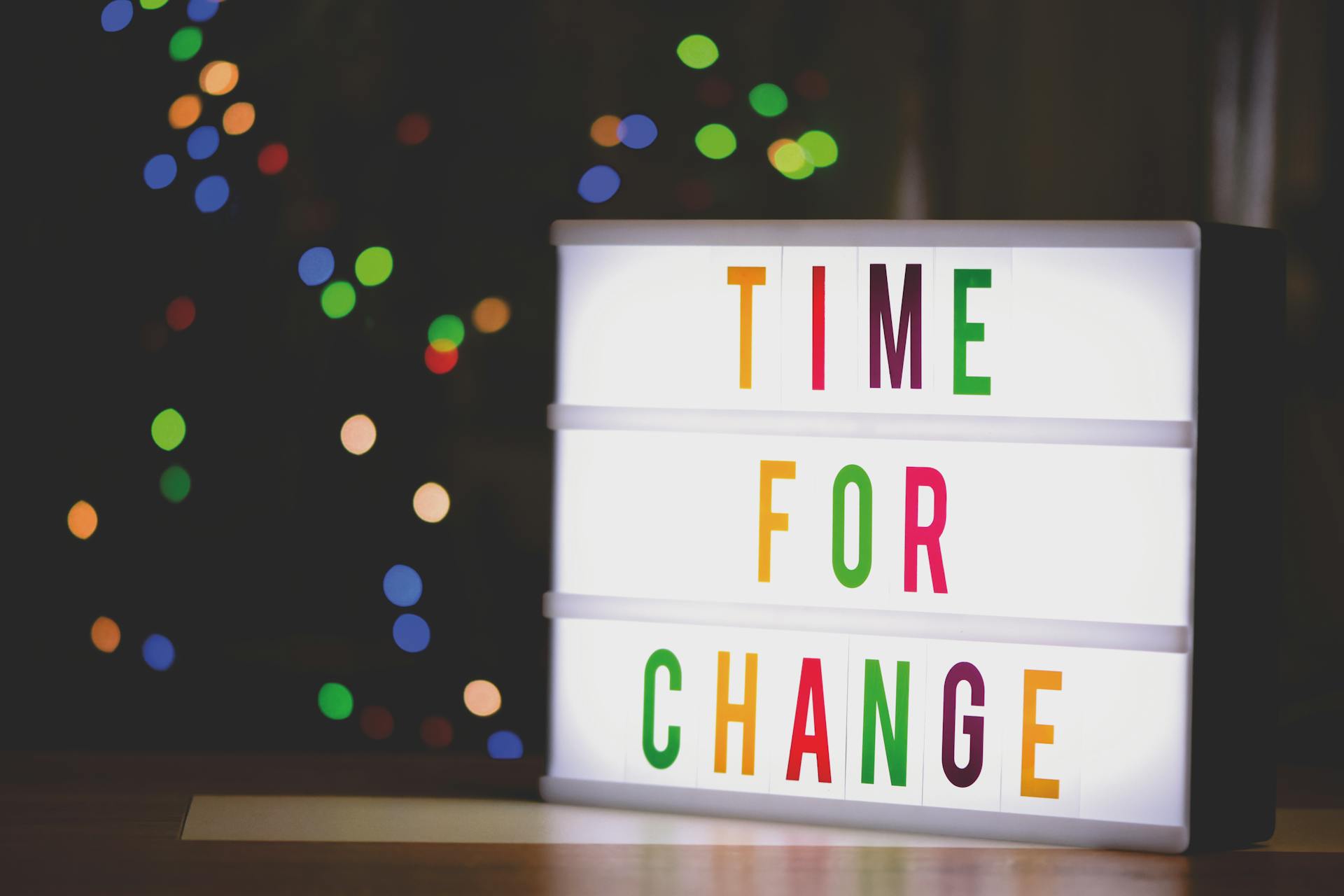 The image is from Pexels.com and shows a light box with the phrase "time for change" spelled out in rainbow lettering. Multicolor lights are seen against a dark backdrop.